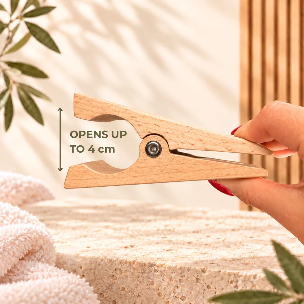 A wooden clothes clip being held open by a hand, with text reading 'OPENS UP TO 4 cm' and a measurement arrow. The scene includes a towel, leaves, and a warm neutral background.