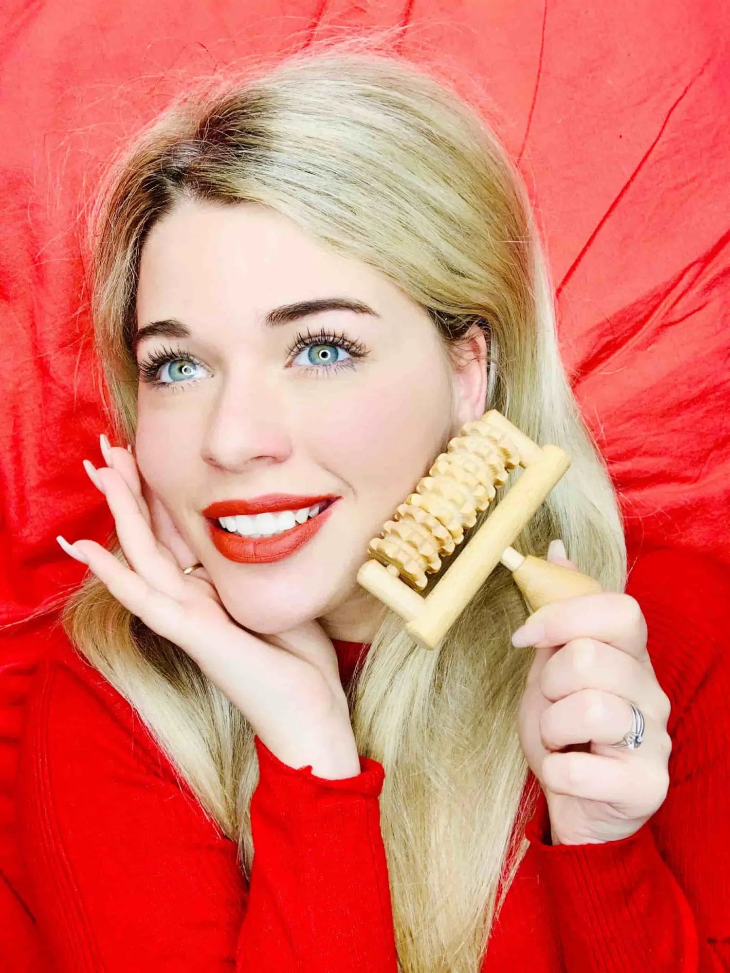 Smiling woman holding a small wooden face massager roller to her cheek on a red background