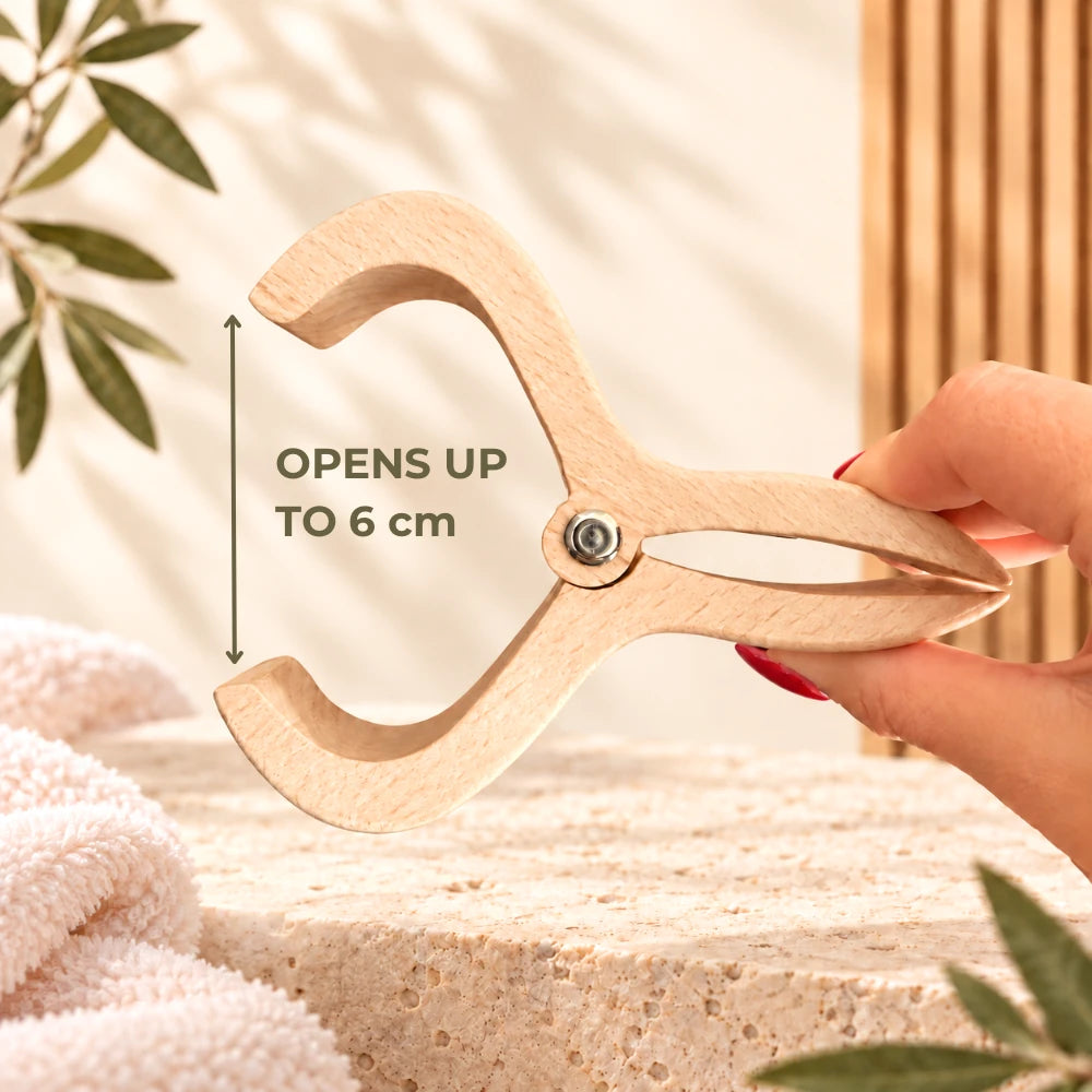 A hand holds a light natural wood hair clamping tool, showing its open width with an arrow overlay that reads 'OPENS UP TO 6 cm'. The background is a warm, neutral spa-like setting with a textured stone surface, soft white towels, and green leaves.
